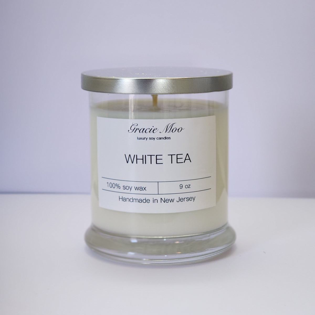 White Tea Status Candle by Gracie Moo Candles, featuring a white wax candle with a simple, elegant design and a lit wick.