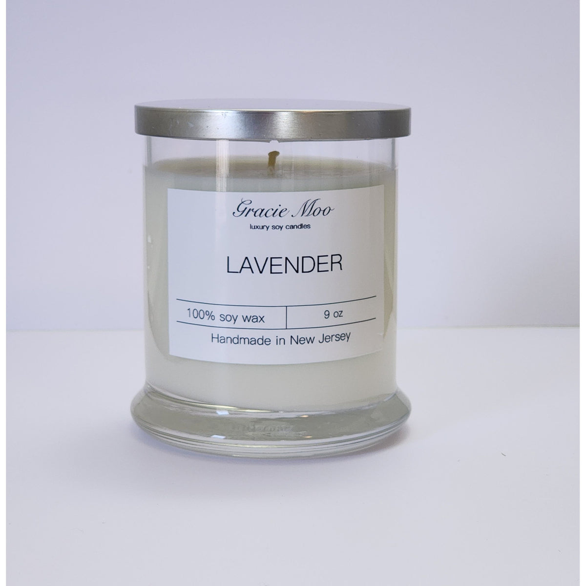 Lavender Status Candle by Gracie Moo Candles with a tall white candle in a glass holder, surrounded by lavender flowers.