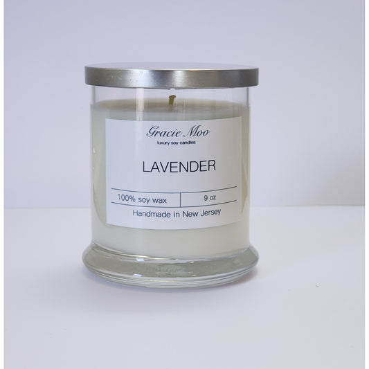 Lavender Status Candle by Gracie Moo Candles with a tall white candle in a glass holder, surrounded by lavender flowers.