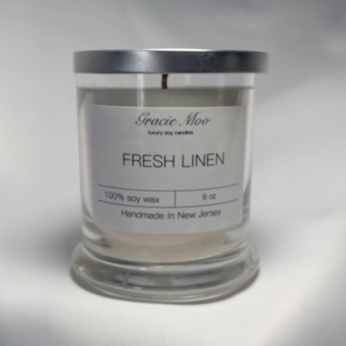 Fresh Linen Status Candle by Gracie Moo Candles, white ceramic candle holder with a lit wick, showcasing a cozy, fresh linen scent.